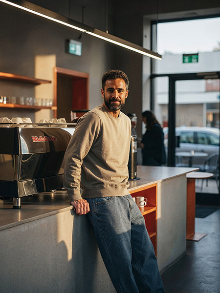 Man leunt tegen de toog van een koffiebar en draagt een sage‑kleurige regular‑fit crewneck uit de Untagged Movement collectie van Texet